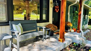 A porch with a wooden bench and a table, a green door, and a garden with flowers and pumpkins.