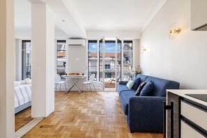 A modern apartment with a herringbone wooden floor, a blue sofa, a dining area with a table and chairs, and a balcony with a view of the city.