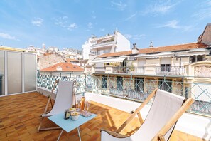 A rooftop terrace with a wooden floor, two lounge chairs, a small table with a bottle and glasses, and a view of the cityscape.