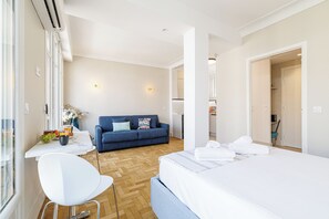A modern hotel room with a bed, a sofa, a dining table with fruits and a vase, and a view of the hallway.