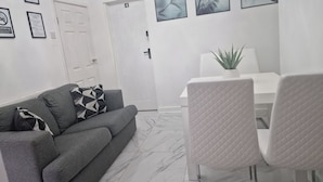 A modern living room with a grey sofa, a white dining table with two chairs, and a door marked with the number 4.