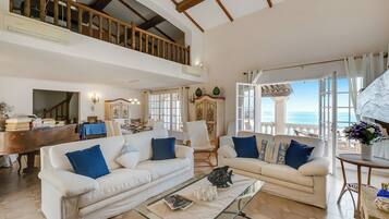 Amplia sala de estar con dos sofás blancos, una mesa de centro de cristal y vistas al mar a través de grandes ventanales.