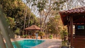 Una piscina con bordo piastrellato, circondata da alberi e una struttura in legno con tetto in tegole.