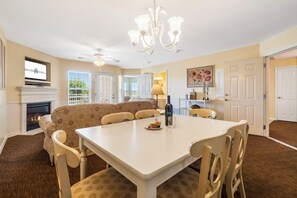 A dining area with a white table, chairs, and a chandelier. There is a fireplace with a TV above it.
