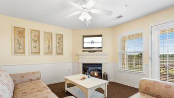 A living room with a fireplace, a ceiling fan, and framed botanical prints on the wall.