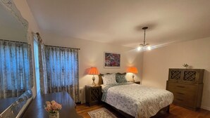 A bedroom with a bed, two bedside tables with lamps, a dresser, a mirror, a window with curtains, and a floral rug.