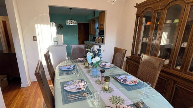 A dining room with a long table set for six, featuring a chandelier, wooden chairs, and a cabinet with glass doors.