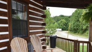 Una terraza de madera con sillas Adirondack y un tapete redondo, un porche cubierto y vistas a un área boscosa.