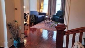 Amplia sala de estar con piso de madera, un sofá azul, un sillón verde y una mesa de centro. Se ve una escalera con barandilla de madera que conduce a otra habitación.