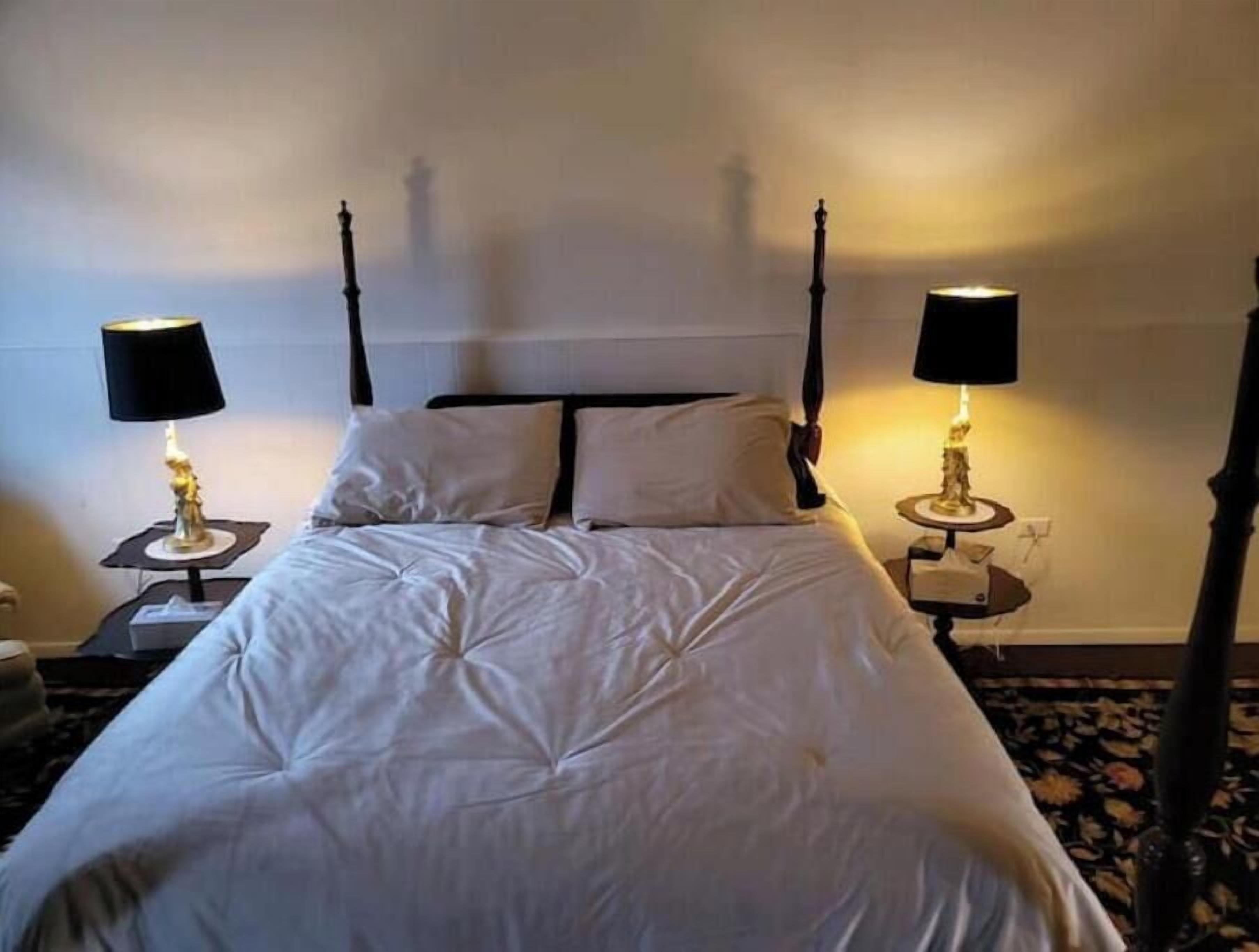 A four-poster bed with white bedding, two bedside tables with lamps, and a plain wall.