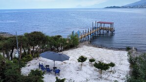 A beach with a wooden pier extending into the water, surrounded by trees and a sandy area with a table and chairs. - House of the Baobas! "Your home in the nature of Atitlán". (San Juan La Laguna)