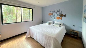 A bedroom with a large window, a bed with white bedding, and a decorative wall piece. - House of the Baobas! "Your home in the nature of Atitlán". (San Juan La Laguna)