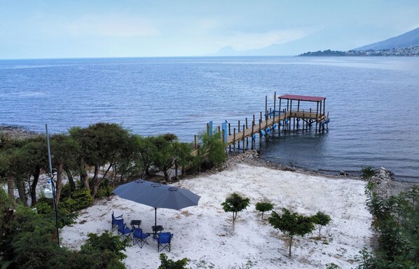 Une plage avec une jetée en bois qui s’avance dans l’eau, entourée d’arbres et d’une zone sablonneuse où se trouvent une table et des chaises.