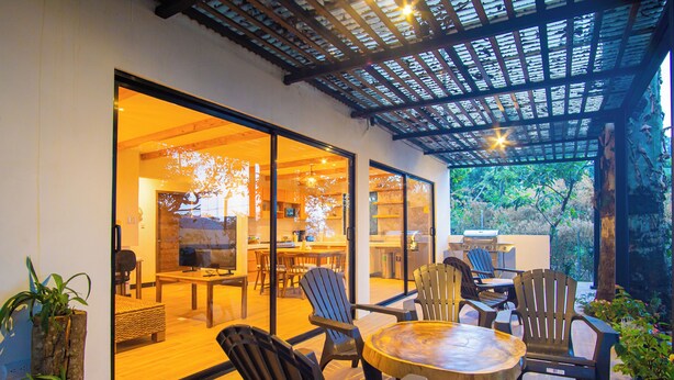 Un espace repas extérieur moderne, avec des chaises en bois et une table ronde, relié à un espace intérieur par une porte vitrée.