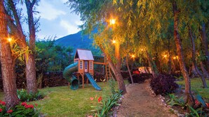 Une cabane de jeu pour enfants avec un toboggan, entourée d’arbres et bordée d’un chemin de jardin.