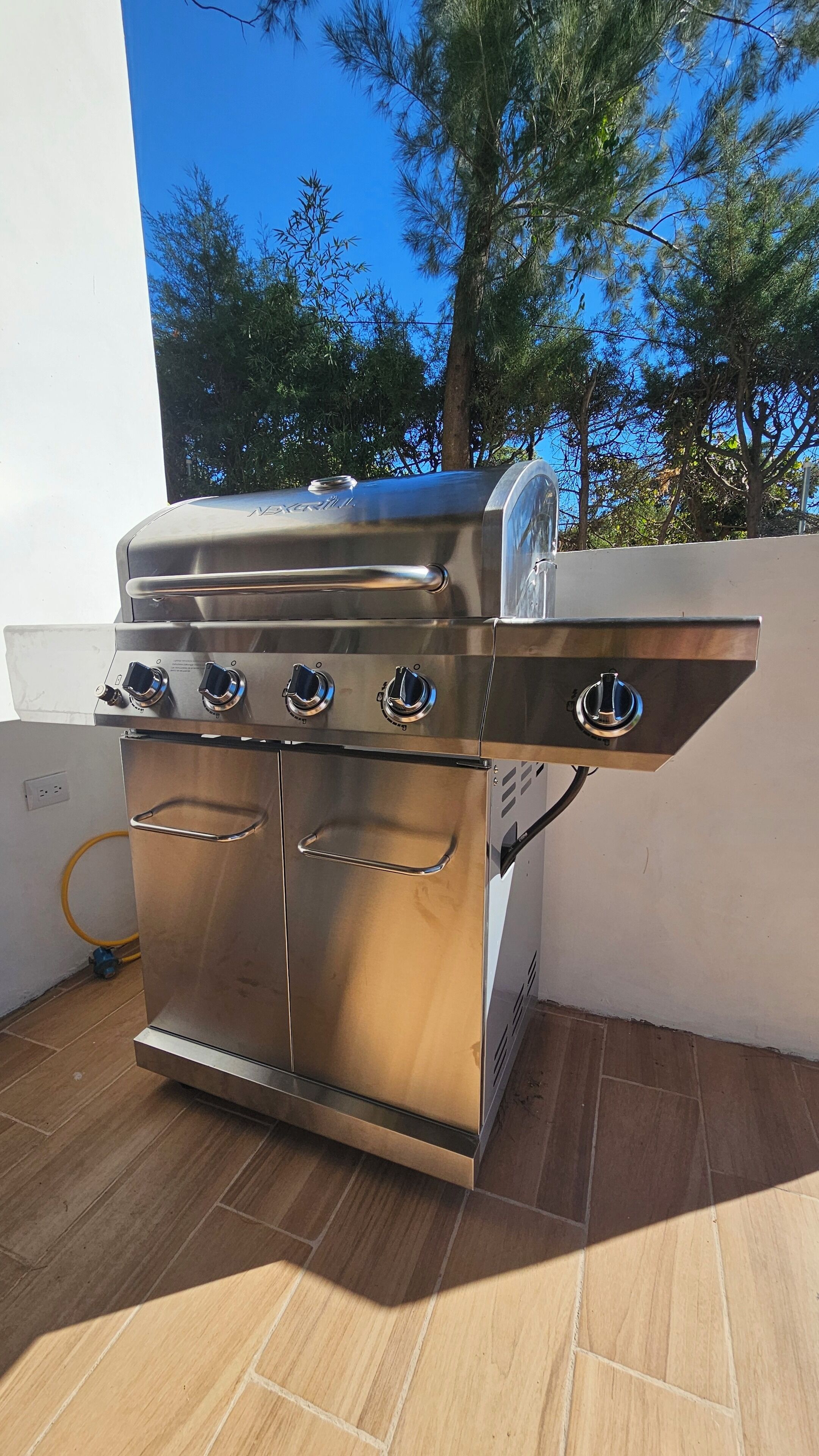 Una parrilla de acero inoxidable con cuatro quemadores y un quemador lateral, ubicada en un patio con baldosas, una pared blanca y árboles al fondo.