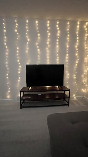 Una habitación con una televisión de pantalla plana sobre un soporte de madera, un sofá gris y luces colgantes en la pared.