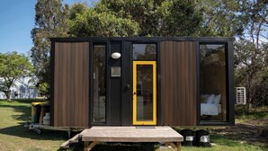 Una cabana moderna i compacta, amb façana de fusta i una porta groga, on es pot veure un llit a través d'una finestra gran.