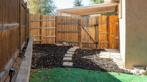 Un patio trasero cercado, con una puerta de madera, un sendero de piedras y una zona de césped artificial verde.