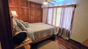 A bedroom with a wooden headboard, a bed with a beige blanket, a bedside table with a lamp, and a window with curtains. - Cozy 3 bedroom House in Quepos center and near Manuel Antonio National Park (Quepos)