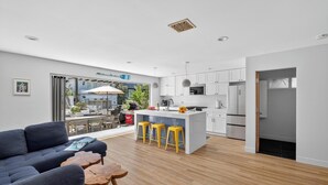 A modern kitchen with white cabinets, a central island with stools, and a dining area with a sofa and wooden stools.