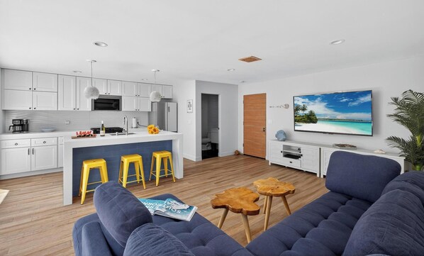 Un espacio habitable moderno con cocina, comedor y un televisor grande que muestra una escena de playa.