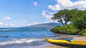 A yellow canoe with "Po'okela" written on it, resting on a sandy beach with a clear blue sky and a tree in the background.