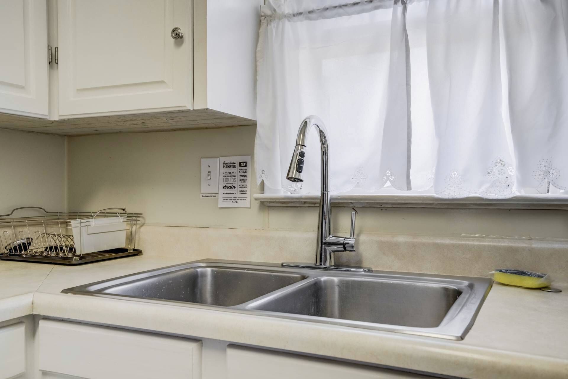 A kitchen with a double stainless steel sink, a window with white curtains, and a white cabinet above the sink.