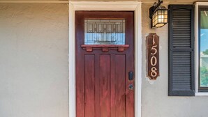 Una puerta roja con una pequeña ventana arriba, una placa con el número de la casa y un farol adosado a la pared.