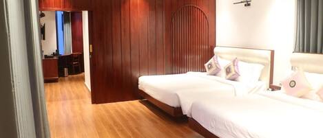 A hotel room with a wooden floor, two beds with white linens, a wooden wardrobe, and a small kitchenette.