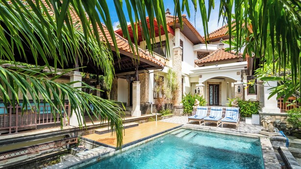 Una piscina con sillas de descanso, rodeada de plantas tropicales y un edificio con tejado de baldosas.