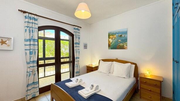 A bedroom with a bed, a nightstand, a lamp, a painting, and a window with curtains. - Rustic and peaceful with pool (Lagos)