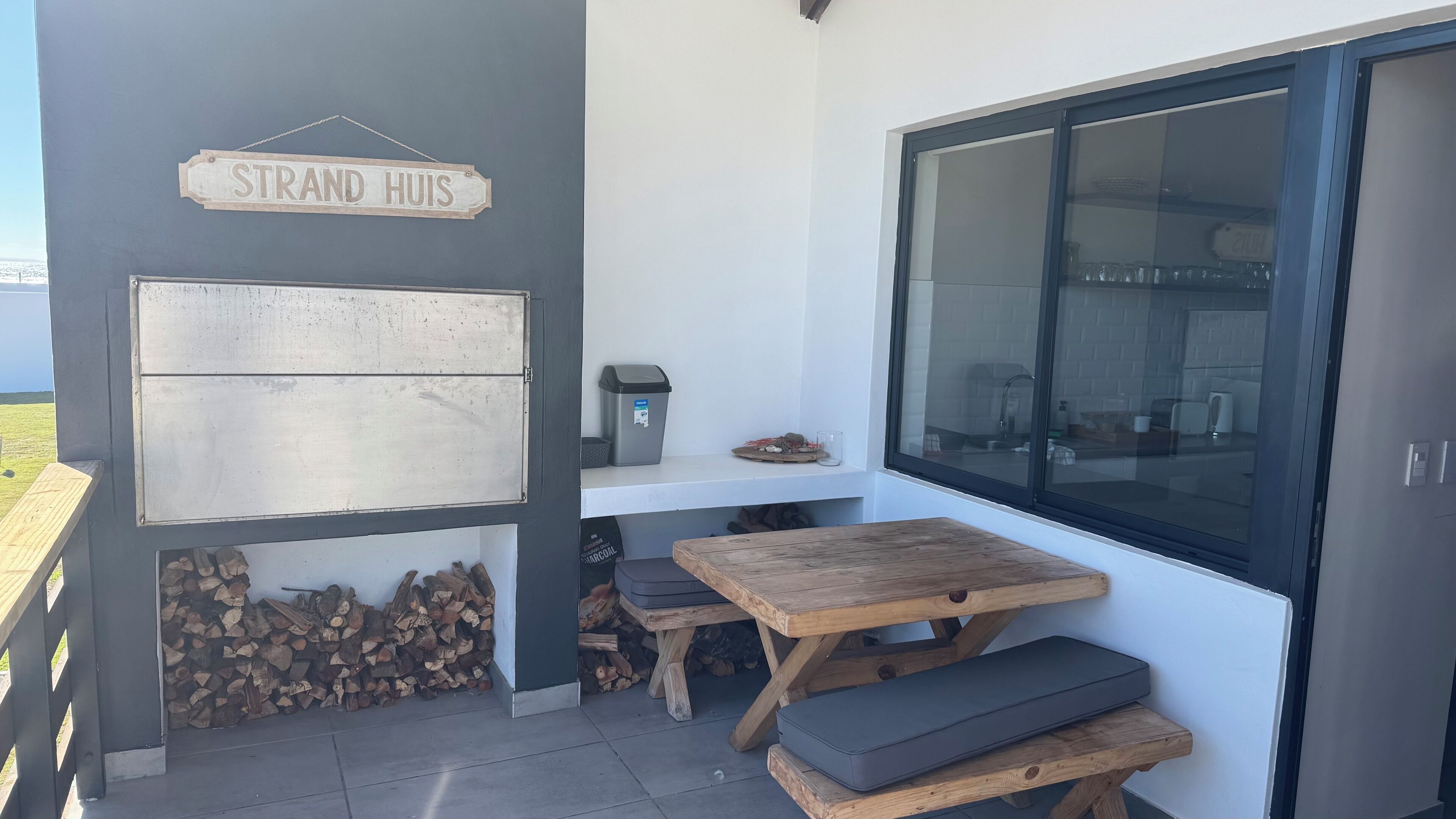 A small outdoor seating area with a wooden table and benches, a trash bin, and a sign reading "STRAND HUIS".