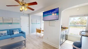 Een moderne woonkamer met een blauwe bank, een eethoek, een keuken met een televisie en een plafondventilator.