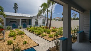 Een modern huis met een overdekte veranda, een pad en een tuin met diverse planten en een blauwe stoel.