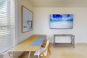 A room with a wooden dining table, white chairs, a small wooden side table, and a mounted TV displaying a beach scene.