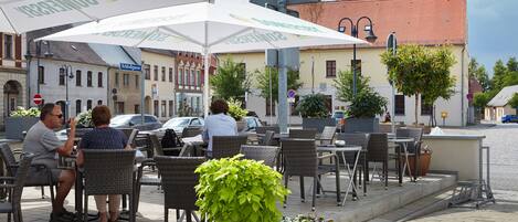 An outdoor cafe with wicker chairs and tables, potted plants, and a building with a red roof in the background.