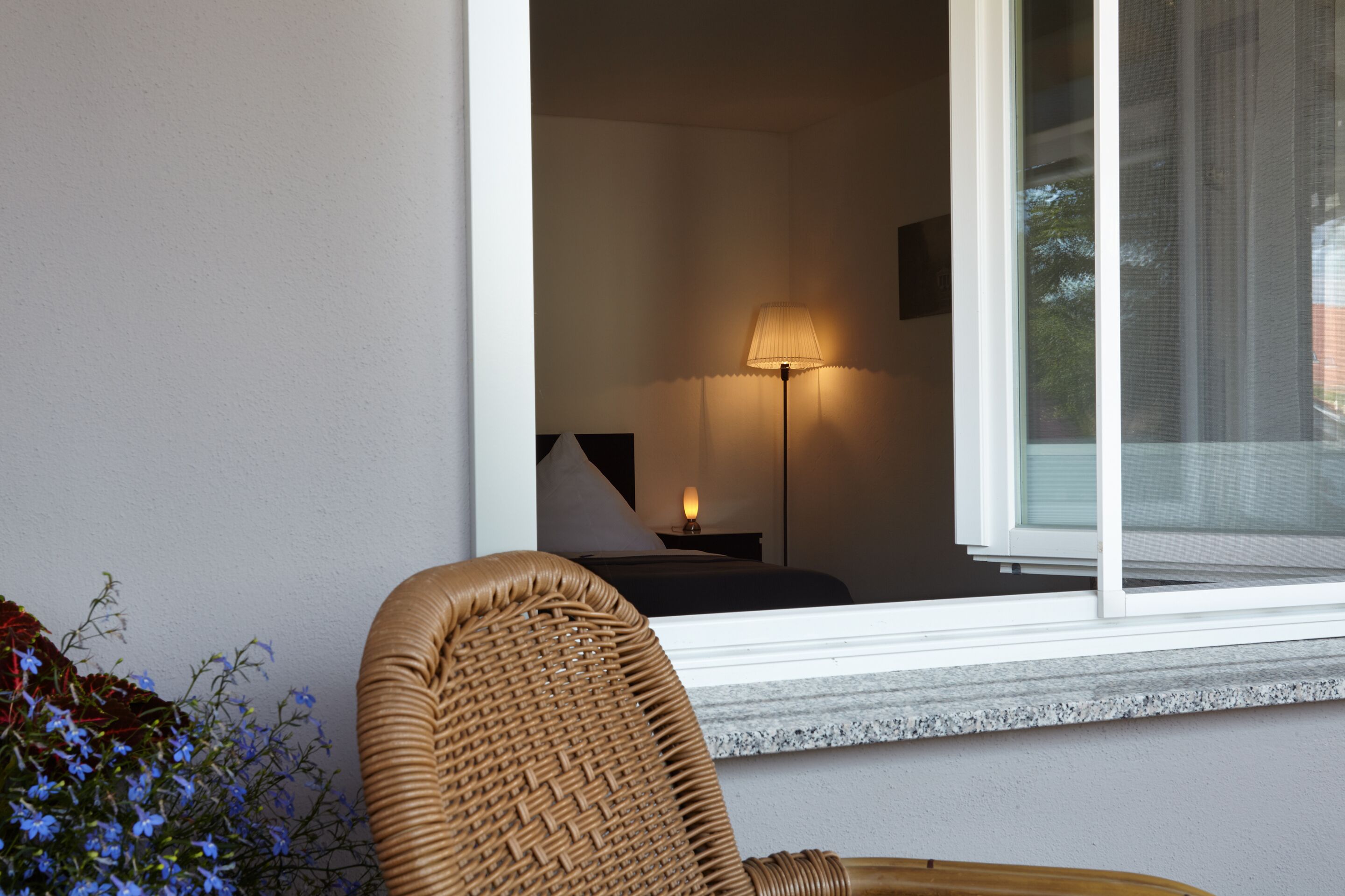 A room with a wicker chair, a bed, a nightstand, and a lamp.