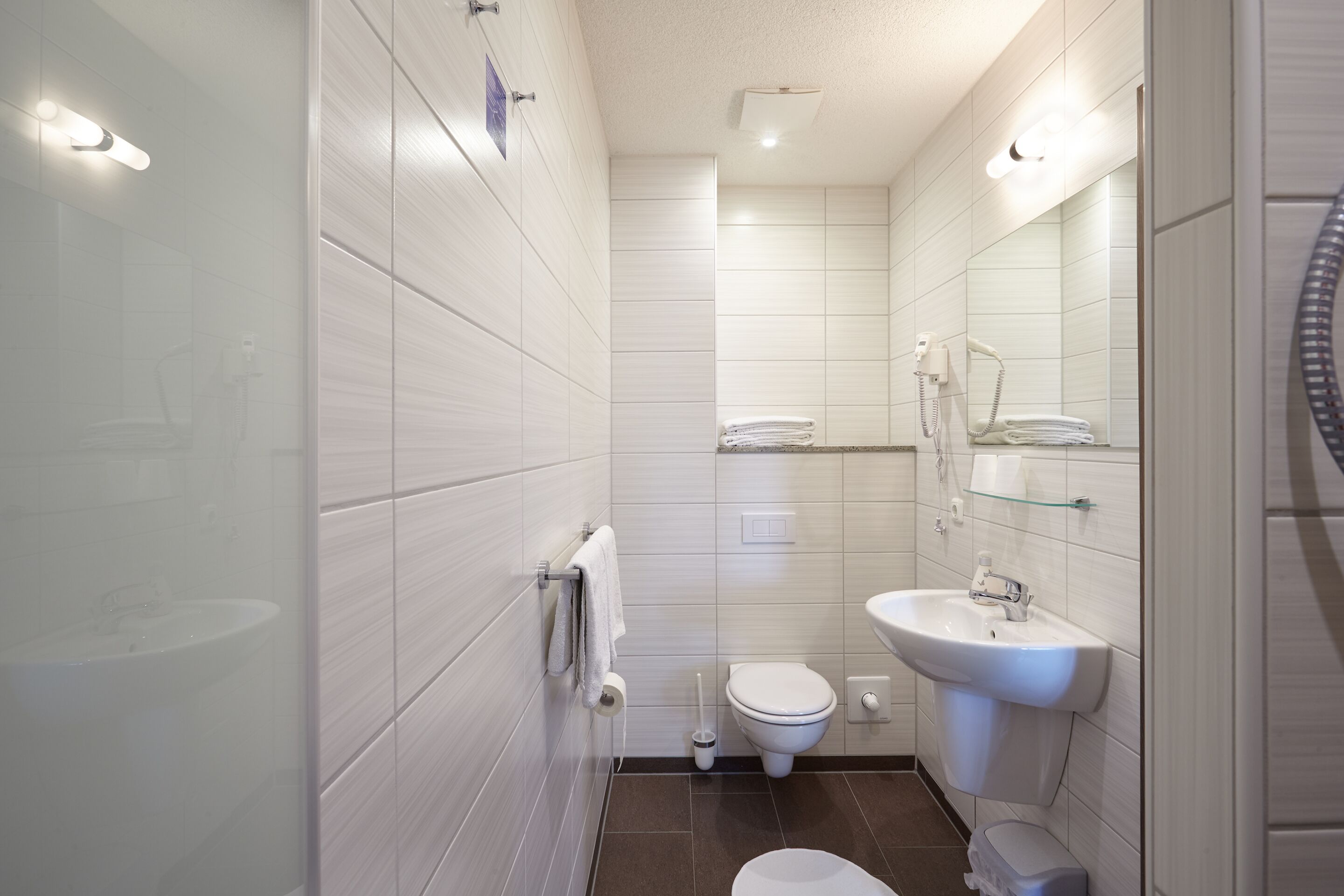 A modern bathroom with white tiles, a toilet, a sink, and a glass shower enclosure.