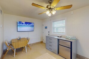 A compact room with a ceiling fan, a flat-screen TV, a small desk with drawers, a chair, and a window with blinds.
