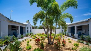 A residential area with two white houses, each with a blue door and windows, surrounded by a garden with various plants and a central tree. - “The Silver Suite” – Steps to Beach, Walk to John’s Pass, Grill,Tickets Included to Top Attractions. (Treasure Island)