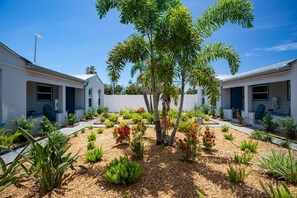 A residential area with two white houses, each with a blue door and windows, surrounded by a garden with various plants and a central tree. - “The Silver Suite” – Steps to Beach, Walk to John’s Pass, Grill,Tickets Included to Top Attractions. (Treasure Island)