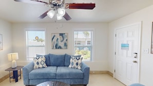 A living room with a blue sofa, a coffee table, a ceiling fan, and two windows with blinds. - “The Silver Suite” – Steps to Beach, Walk to John’s Pass, Grill,Tickets Included to Top Attractions. (Treasure Island)