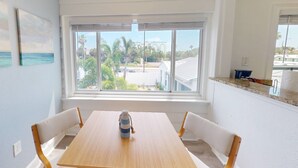Un comedor con una mesa y sillas, una ventana con vistas a palmeras y edificios, y una encimera de cocina con un recipiente azul.