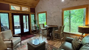 Ein geräumiges Wohnzimmer mit hoher Decke, Holzbalken, einer Marmorwand und großen Fenstern mit Blick ins Freie.