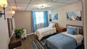 A modern bedroom with two beds, a large window with blue curtains, a ceiling with a grid pattern, and a painting on the wall.