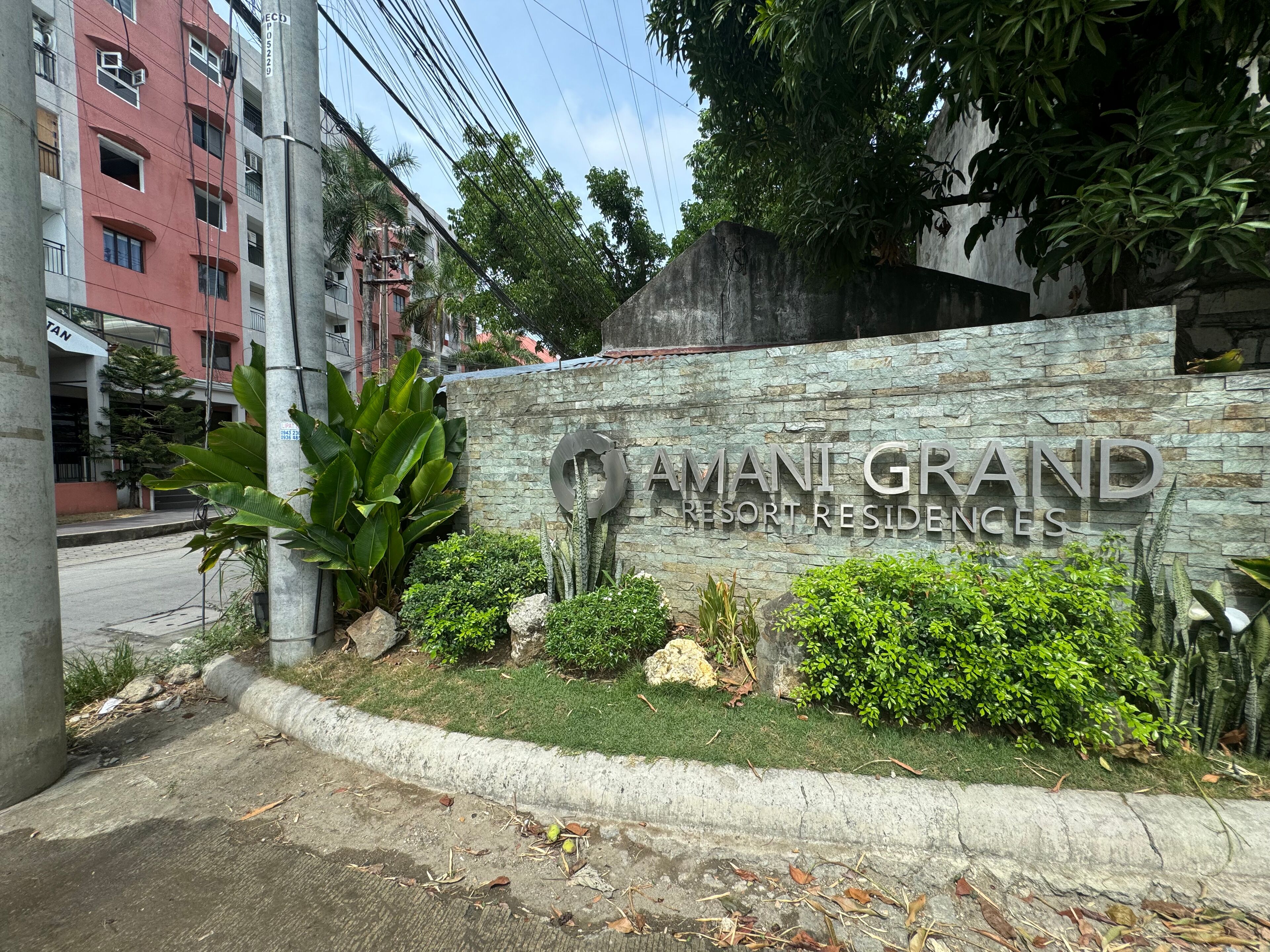 A sign for Amani Grand Resort Residences with a stone wall and greenery.