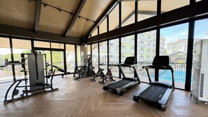 Een sportschool met trainingsapparatuur, grote ramen en uitzicht op gebouwen en een zwembad.