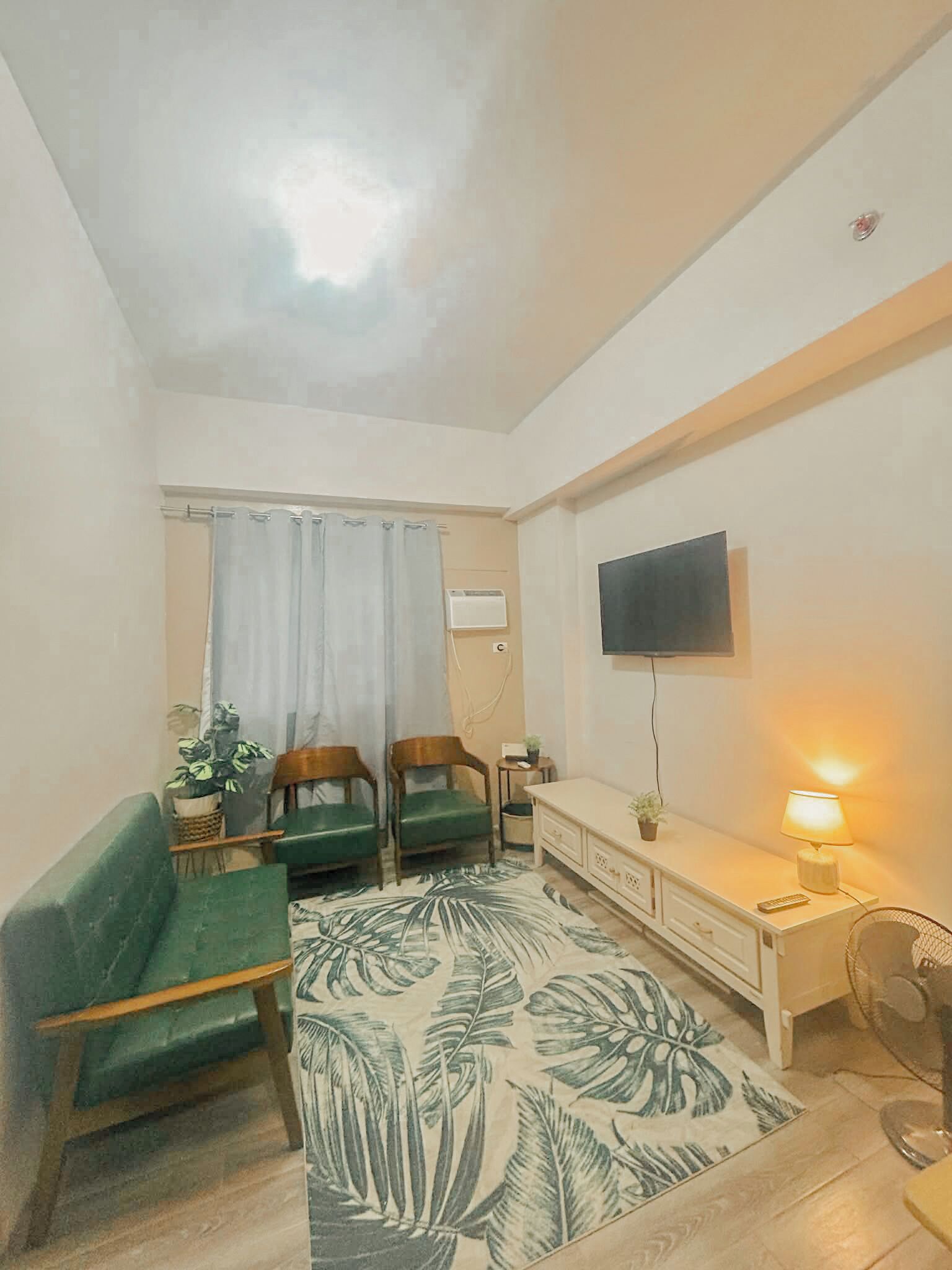 A small, well-lit room with a flat-screen TV mounted on the wall, a green sofa, two wooden chairs, a small table with a lamp, and a patterned rug.