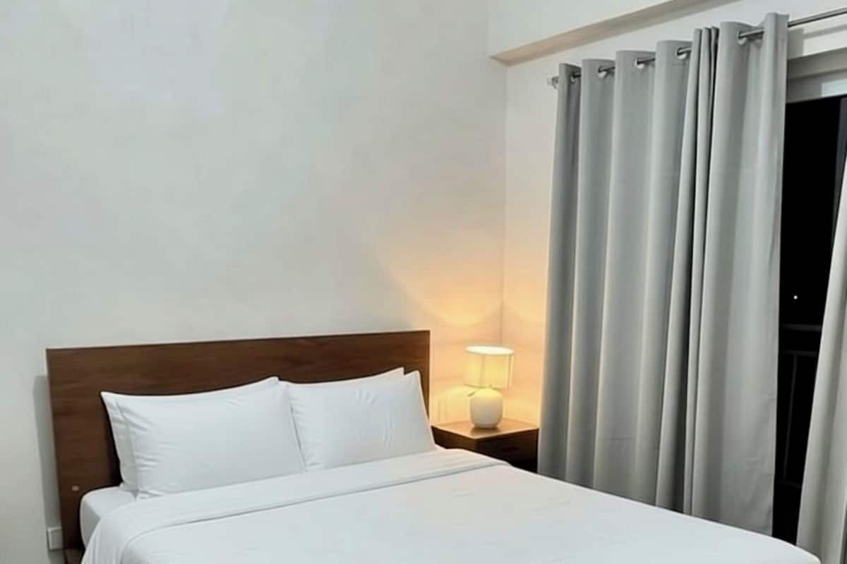 Un lit simple avec une tête de lit en bois, une lampe de chevet et une vue sur une fenêtre avec des rideaux.
